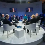 Uma foto do World Economic Forum com diversas pessoas sentadas em uma sala com formato de círculo e com um palco central (em branco) e com 6 pessoas sentadas e conversando.