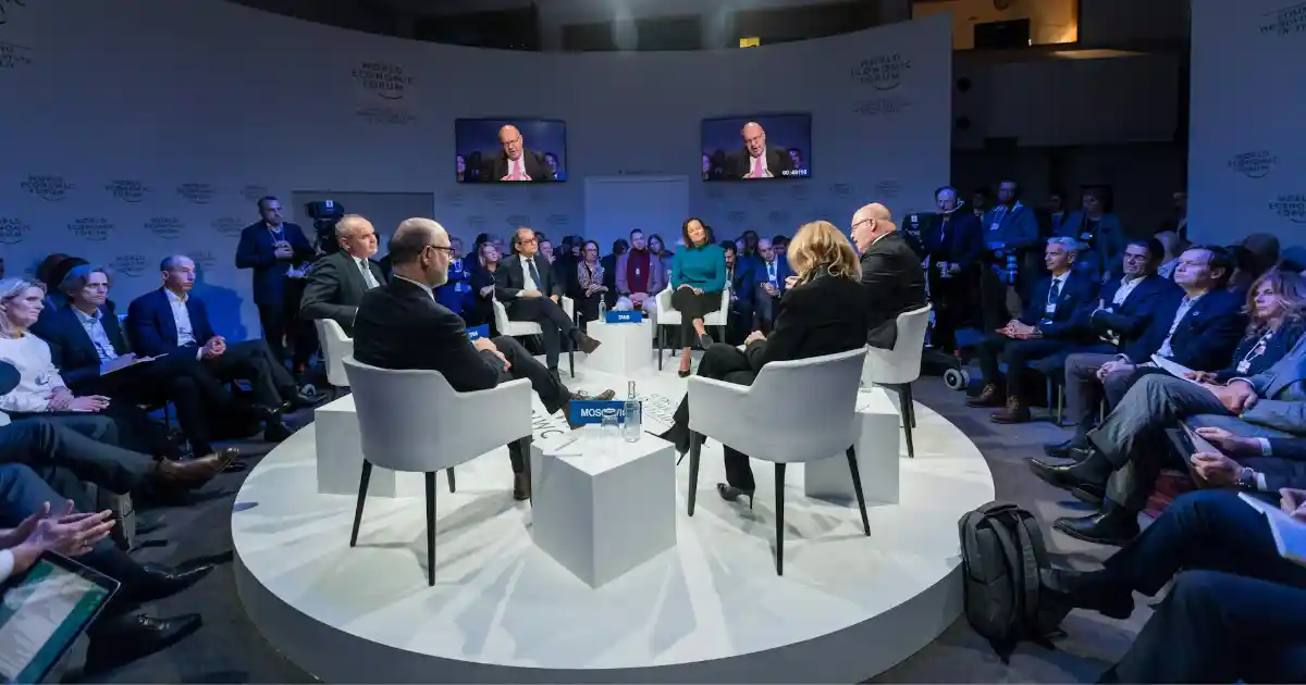 Uma foto do World Economic Forum com diversas pessoas sentadas em uma sala com formato de círculo e com um palco central (em branco) e com 6 pessoas sentadas e conversando.