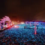 Foto da vista do Palco Mundo do Rock in Rio a partir da tirolesa do evento. Há diversas pessoas no festival e a foto está bem iluminada com bastante luz azul