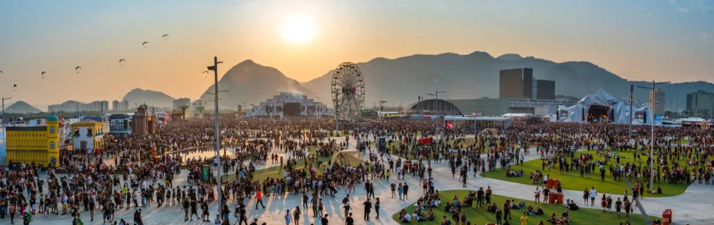 Foto panorâmica do Rock in Rio com várias pessoas, montanhas ao fundo junto com o pôr do sol e a roda gigante em destaque.