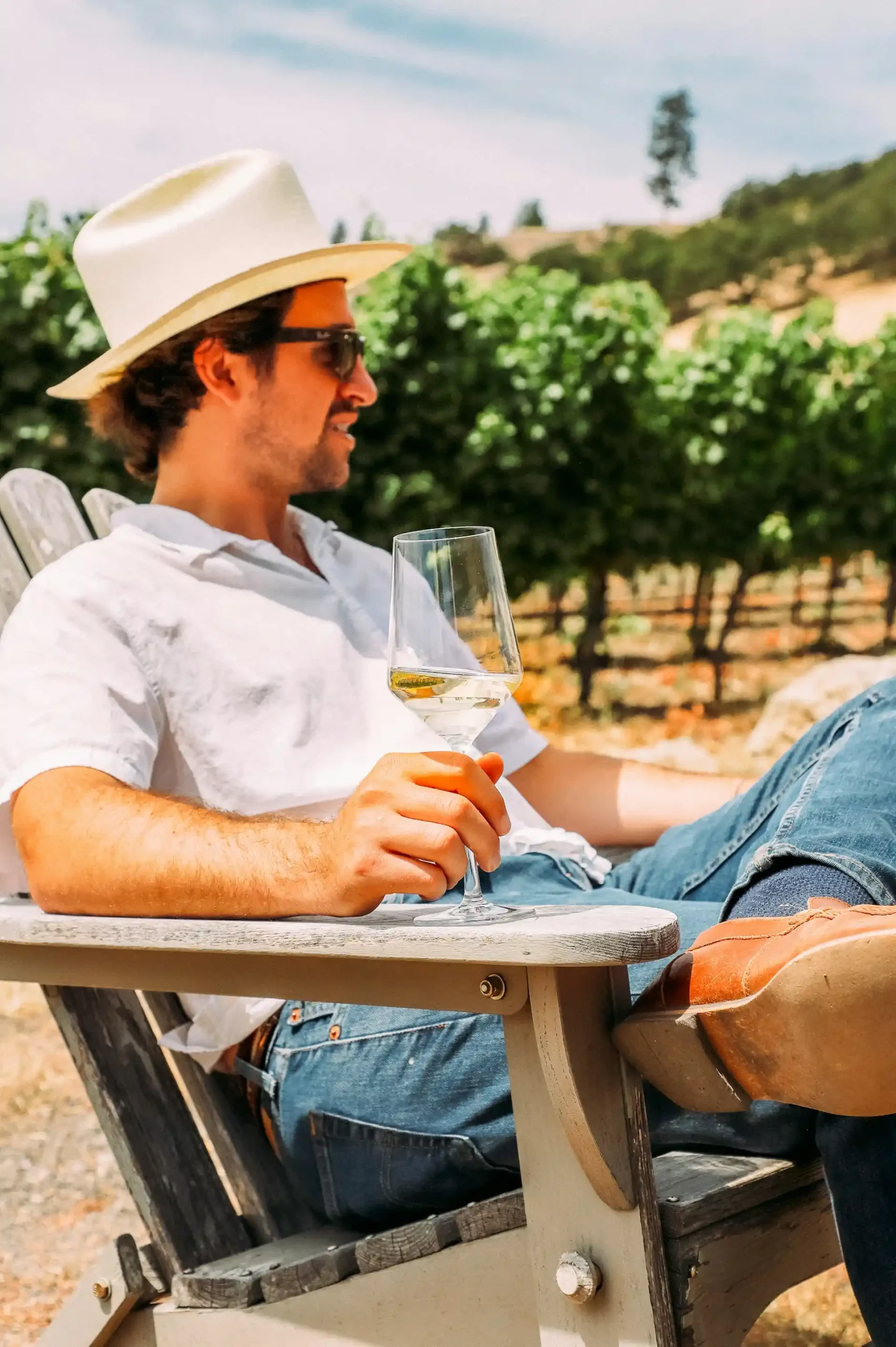 Homem branco com um chapéu branco, camisa branca e calça jeans azul, sentado em uma cadeira de madeira, com uma taça de vinho branco na mão. Ao fundo, um vinhedo