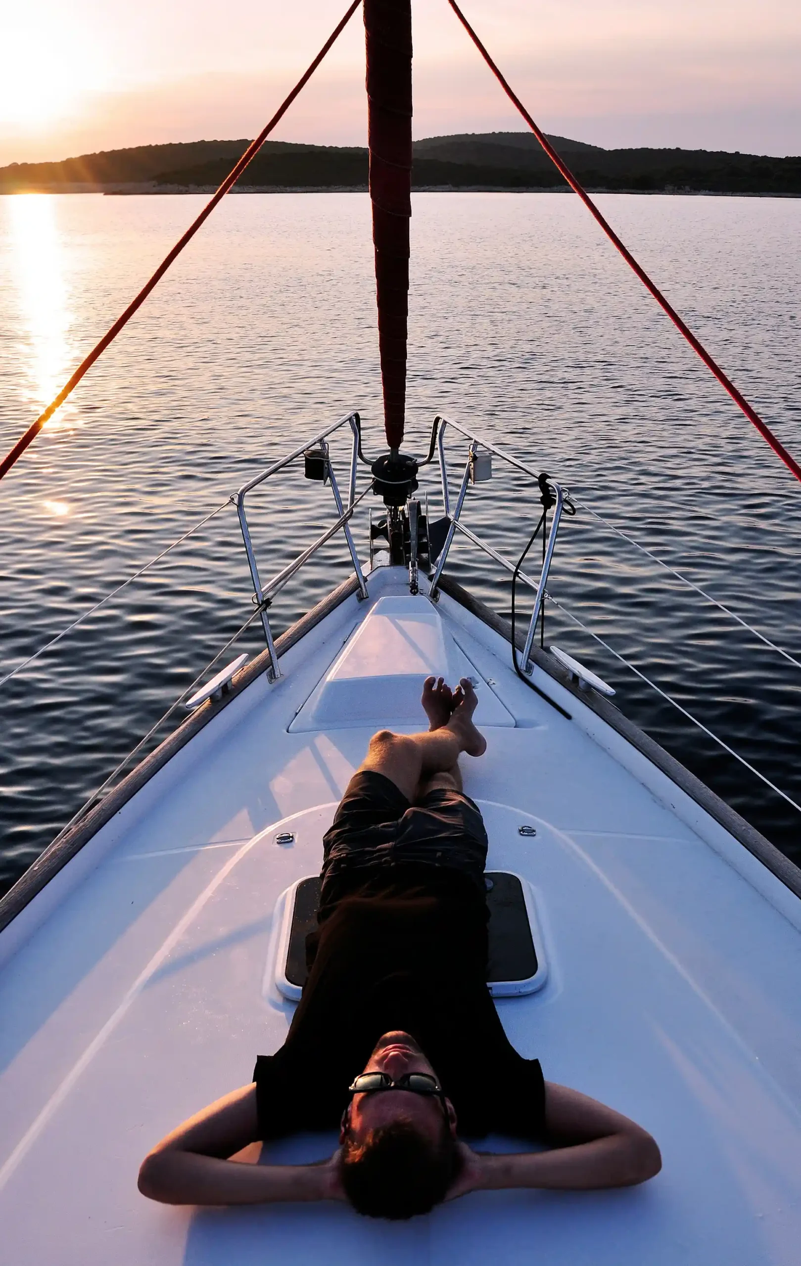 Homem deitado em um barco em alto mar com vista para montanhas ao fundo.