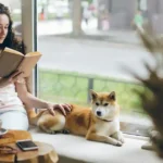 Uma mulher em uma cafeteria lendo um livro com o seu cachorro ao lado