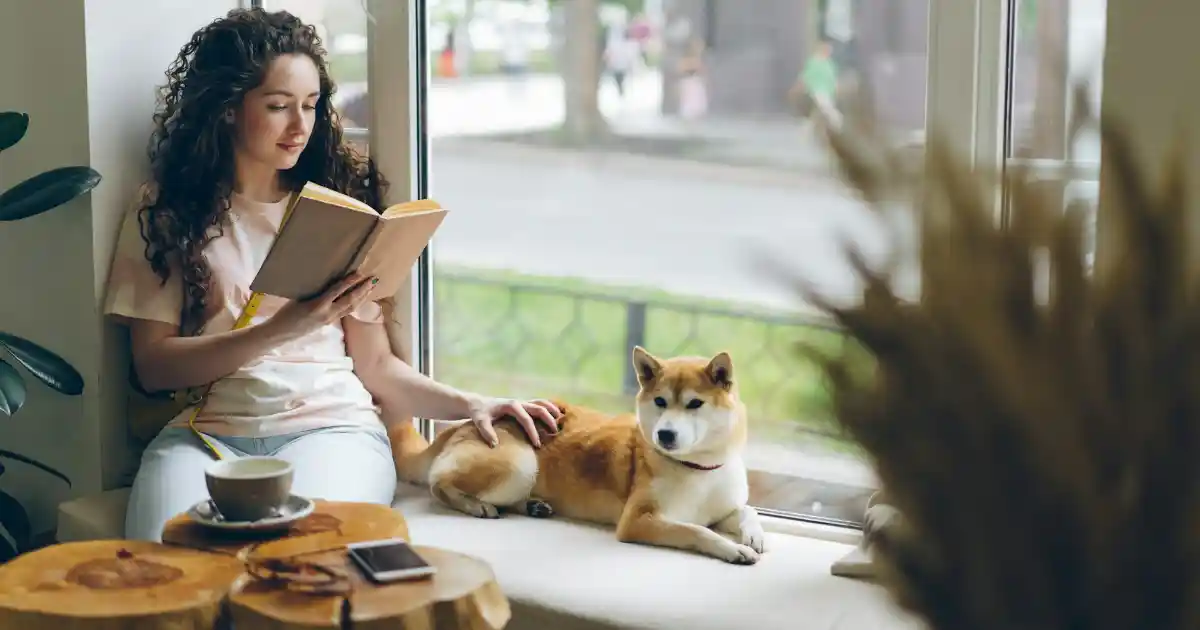 Uma mulher em uma cafeteria lendo um livro com o seu cachorro ao lado